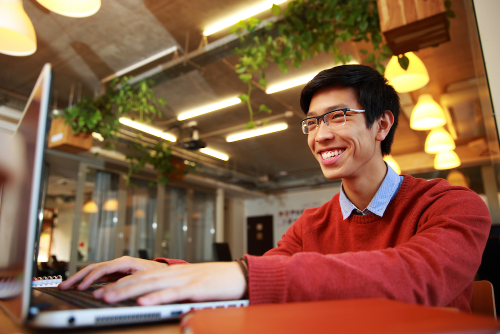 Cheerful Asian man wearing glasses typing on a laptop in a cafe