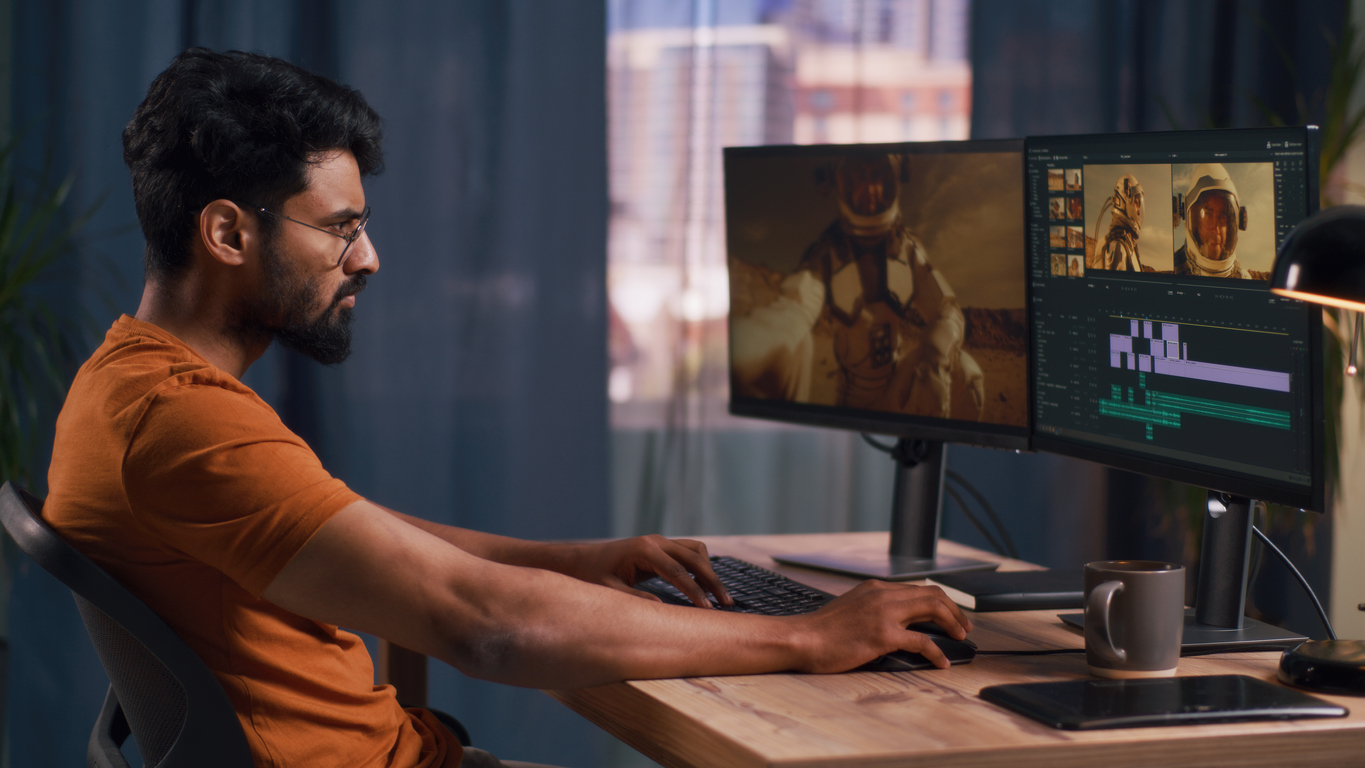 Side portrait of a man using a keyboard shortcut to edit a video on his desktop with extra screen monitor on the side.