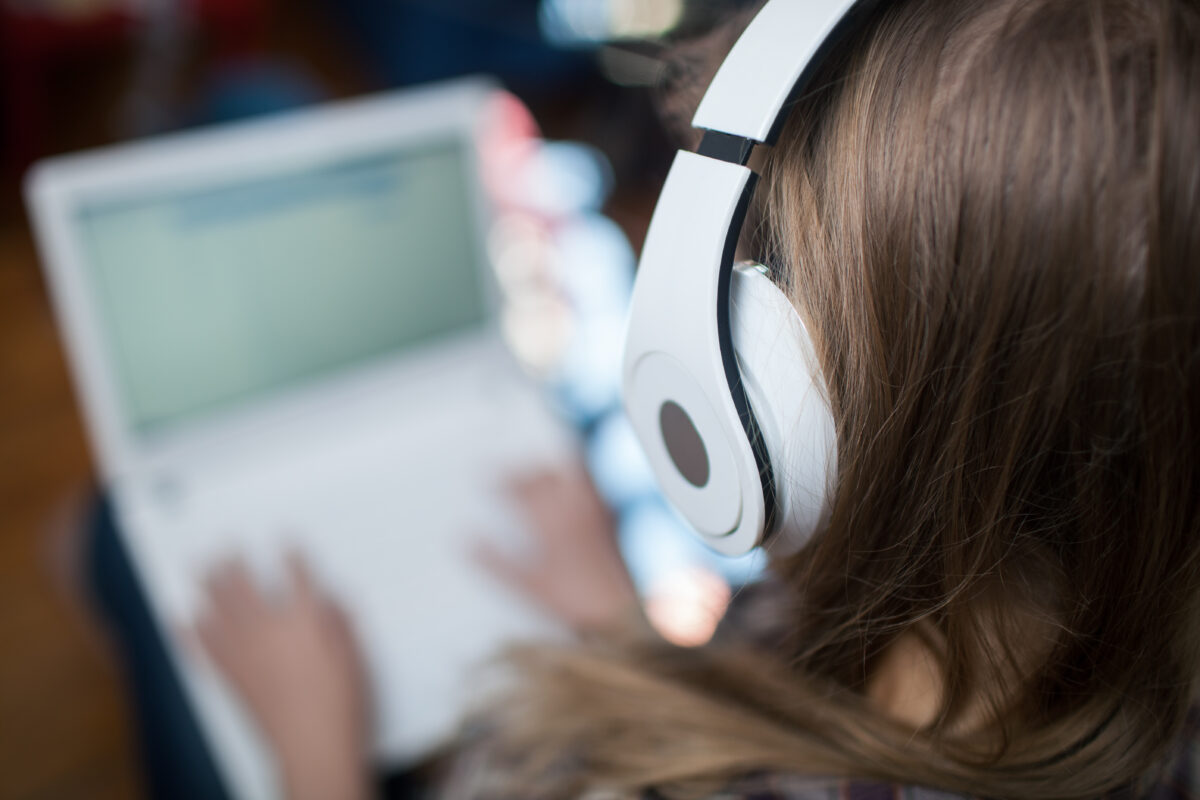 Image taken over shoulder of woman wearing headphones and working on a laptop.